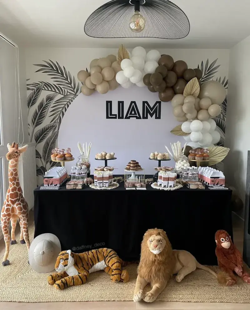 Anniversaire d'enfant sur le thème safari : arche de ballons beige, feuilles de palmier, table sucrée et peluches d'animaux