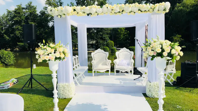 Cérémonie de mariage en extérieur : pergola drapée de blanc, fauteuils des mariés, allée blanche et compositions de fleurs blanches